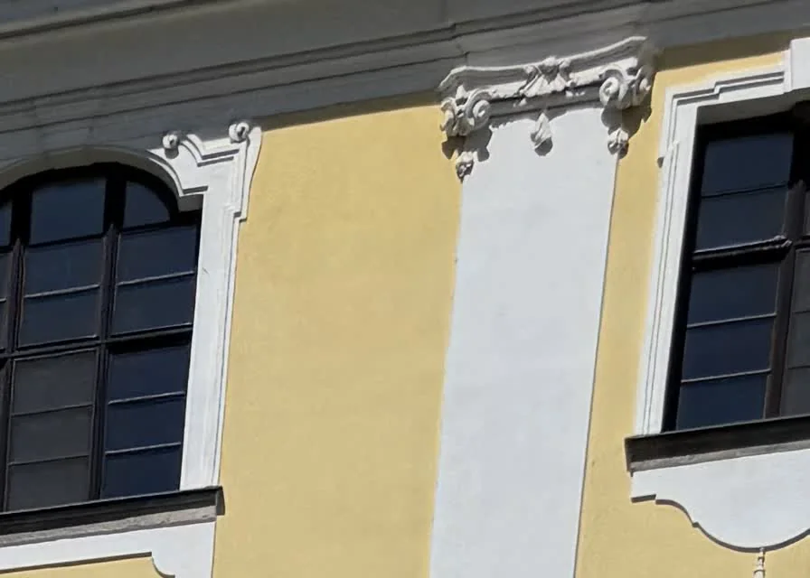 Detailní snímek z fotogrammetrického sběru na historické fasádě: žlutá omítka mezi bílými lizénami, ozdobná štuková hlavice a dvě vysoká okna s obloukovým záklenkem a tmavými rámy.